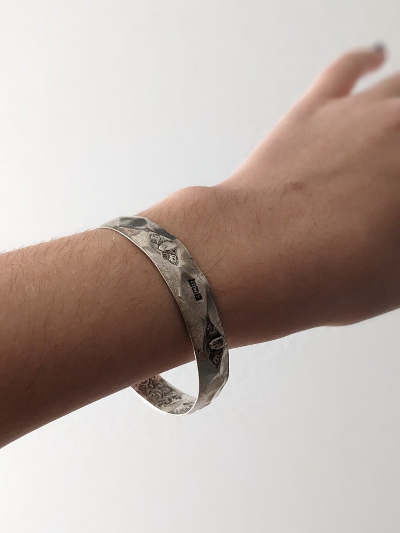 May include: A silver bracelet with a hammered finish and intricate engraved designs. The bracelet has a hallmark that reads "925".