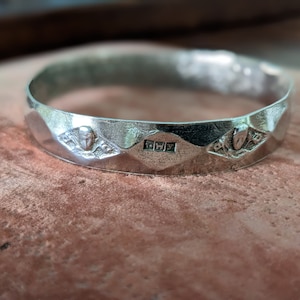May include: A silver bangle bracelet with a faceted design and three decorative details. The center detail is a rectangular stamp with the letters "FWY".