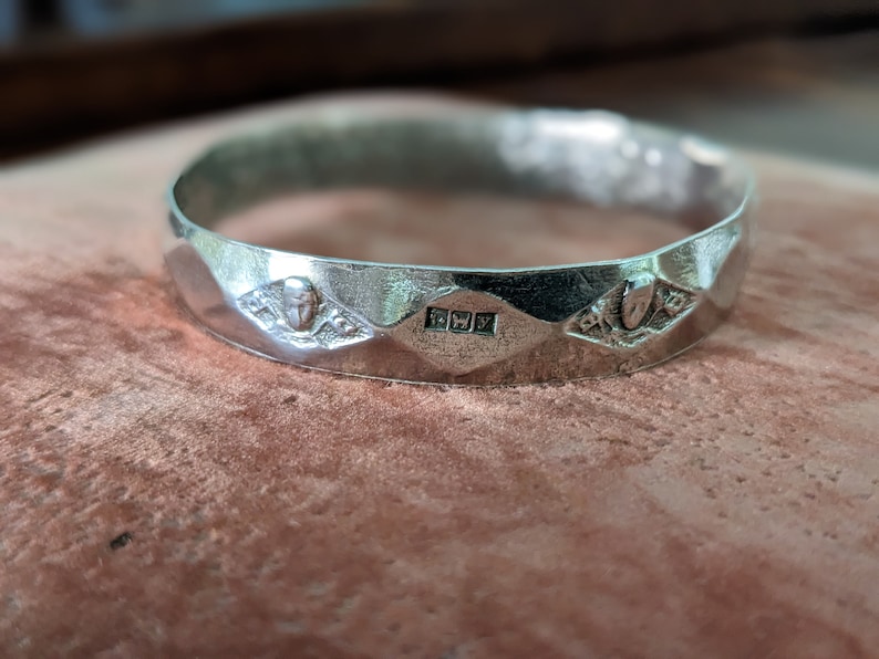 May include: A silver bangle bracelet with a faceted design and three decorative details. The center detail is a rectangular stamp with the letters "FWY".