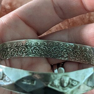 May include: Silver bangle bracelet with a decorative, engraved floral pattern. The bracelet is open and has a smooth, polished finish.