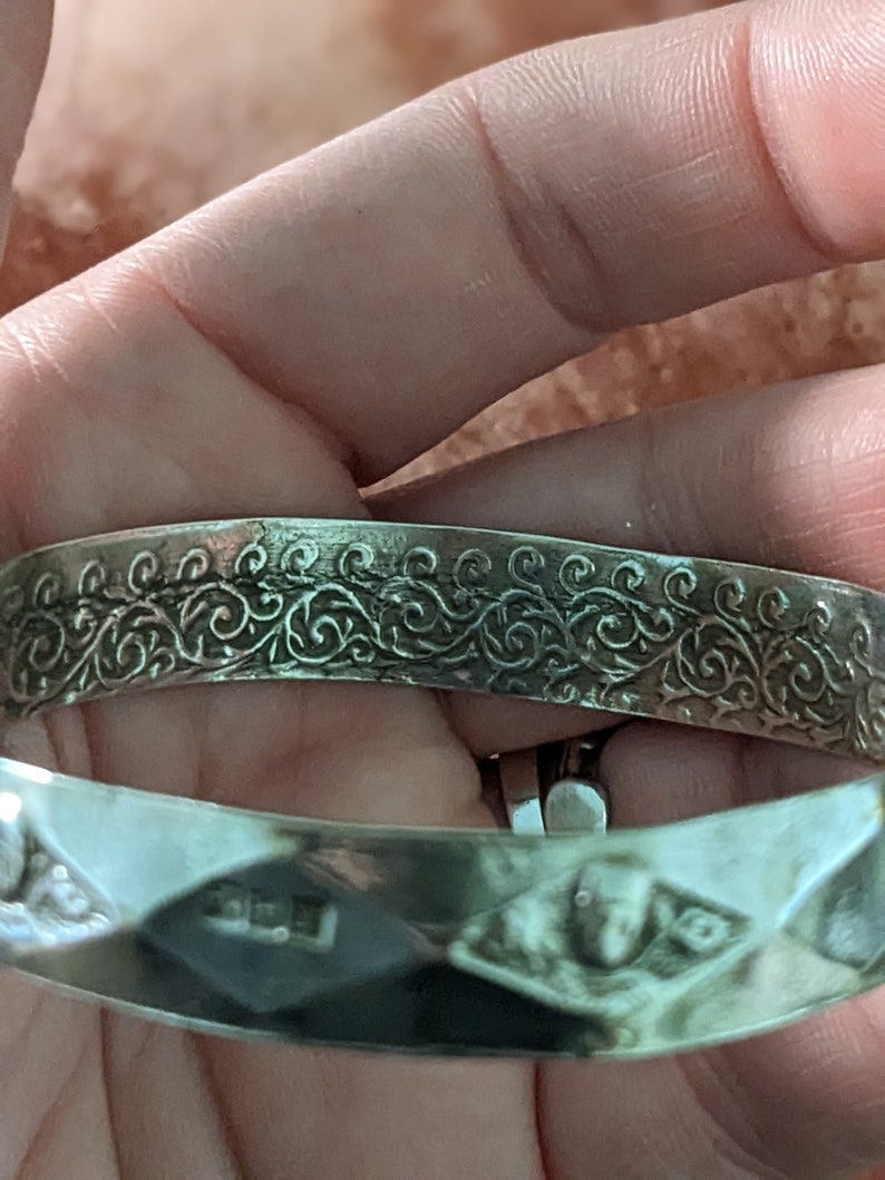 May include: Silver bangle bracelet with a decorative, engraved floral pattern. The bracelet is open and has a smooth, polished finish.