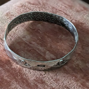 May include: A silver bangle bracelet with an intricate floral design and a hallmark on the bottom. The bracelet is on a pink and brown textured surface.