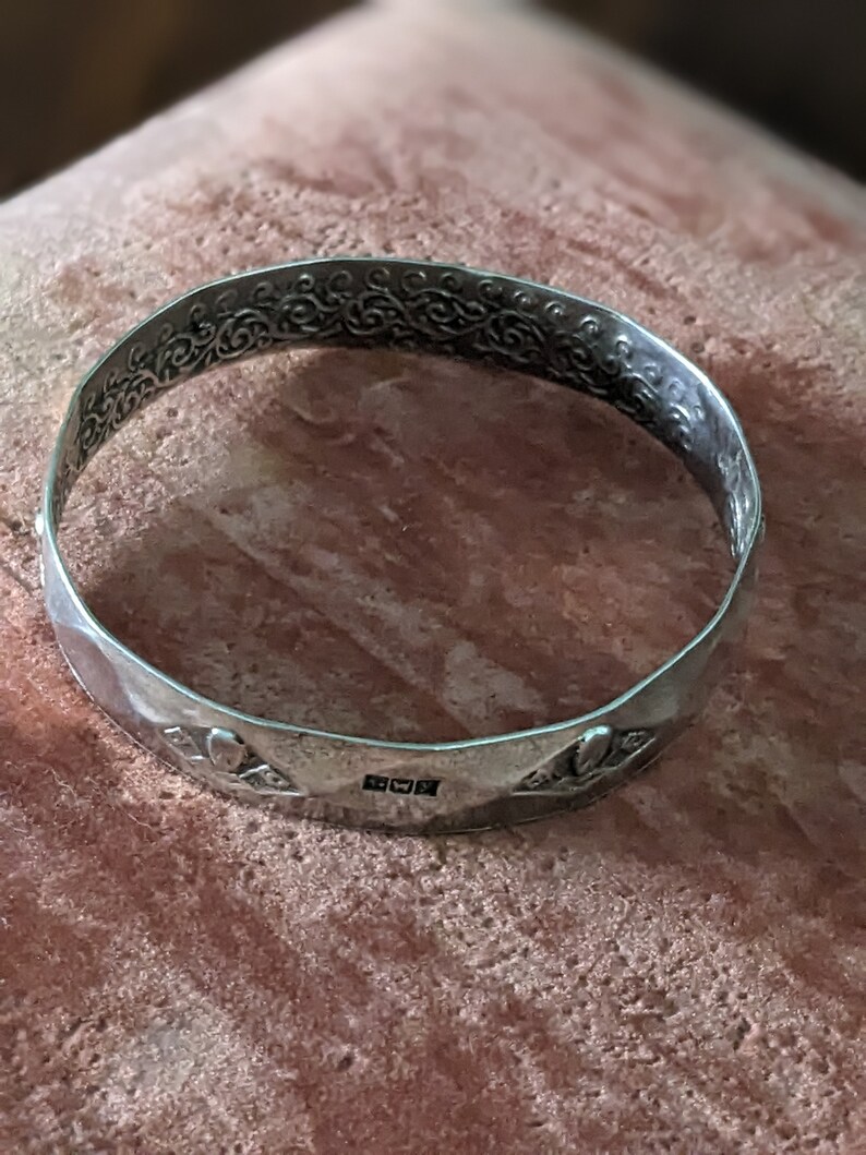 May include: A silver bangle bracelet with an intricate floral design and a hallmark on the bottom. The bracelet is on a pink and brown textured surface.