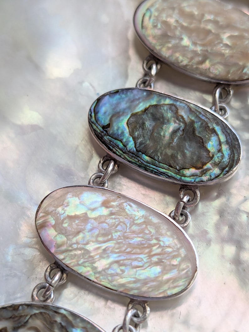 May include: A silver bracelet with oval-shaped abalone shell charms. The shells have iridescent colors of blue, green, and purple.