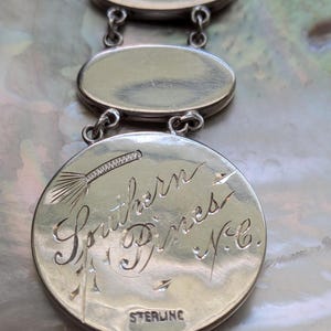 May include: Close-up of a silver charm bracelet with three oval charms. The bottom charm has an inscription that reads "Southern Pines, N.C." and "STERLING".