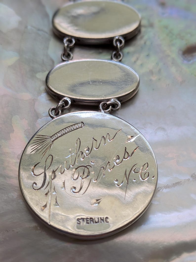 May include: Close-up of a silver charm bracelet with three oval charms. The bottom charm has an inscription that reads "Southern Pines, N.C." and "STERLING".