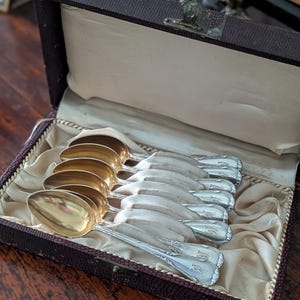 May include: A collection of antique silver spoons with gold-plated bowls, presented in a brown velvet-lined box. The spoons are arranged on a cream satin lining, with decorative handles. The box is secured with a metal clasp, showcasing a vintage aesthetic.