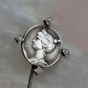 May include: Silver stick pin with a round, silver medallion featuring a profile of a woman with a crown. The medallion is surrounded by four small, clear stones.