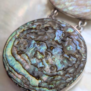 May include: A silver necklace with two round abalone shell pendants. The larger pendant has a swirling pattern of blue, green, and brown.