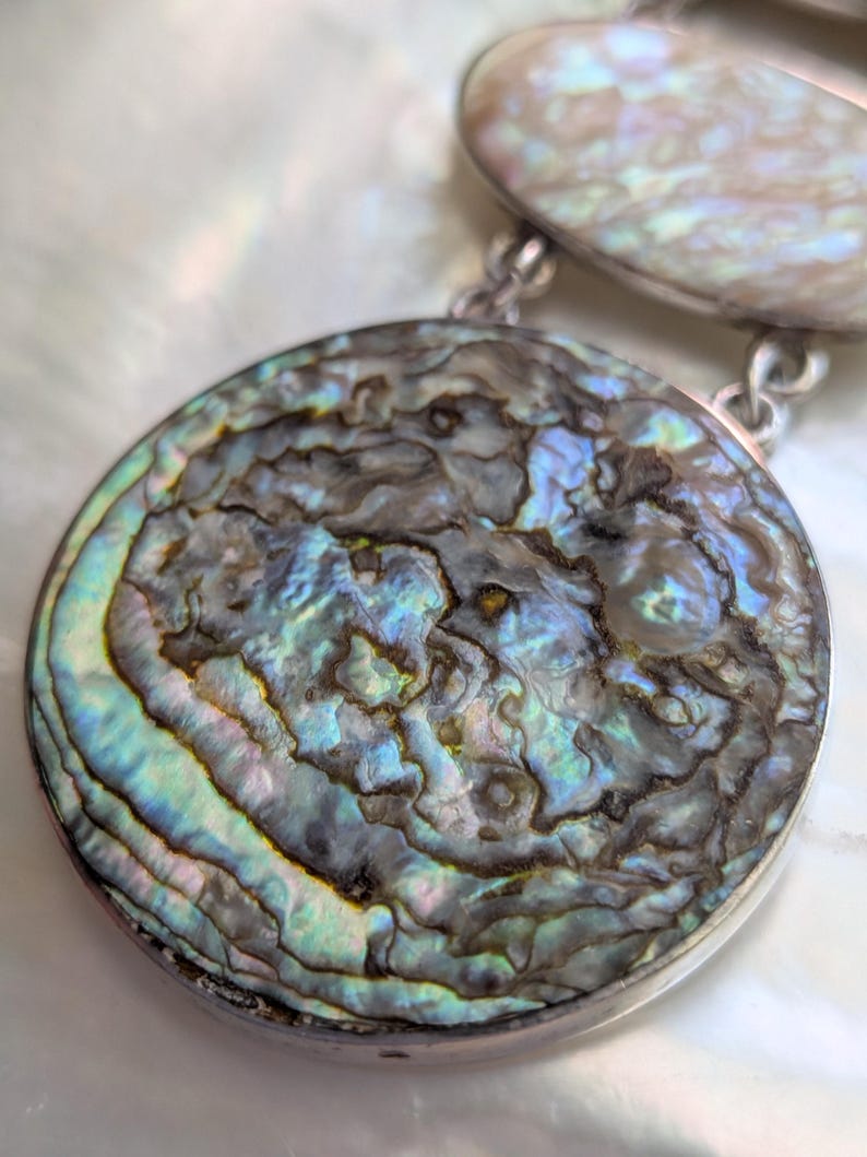 May include: A silver necklace with two round abalone shell pendants. The larger pendant has a swirling pattern of blue, green, and brown.