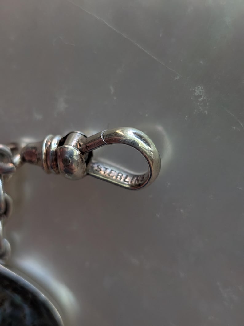 May include: Close-up of a silver pocket watch chain with a clasp that reads "STERLING".
