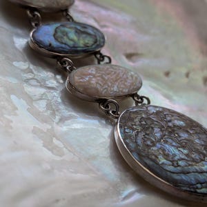 May include: A silver chain necklace with oval-shaped abalone shell pendants. The pendants have a iridescent blue, green, and brown color.
