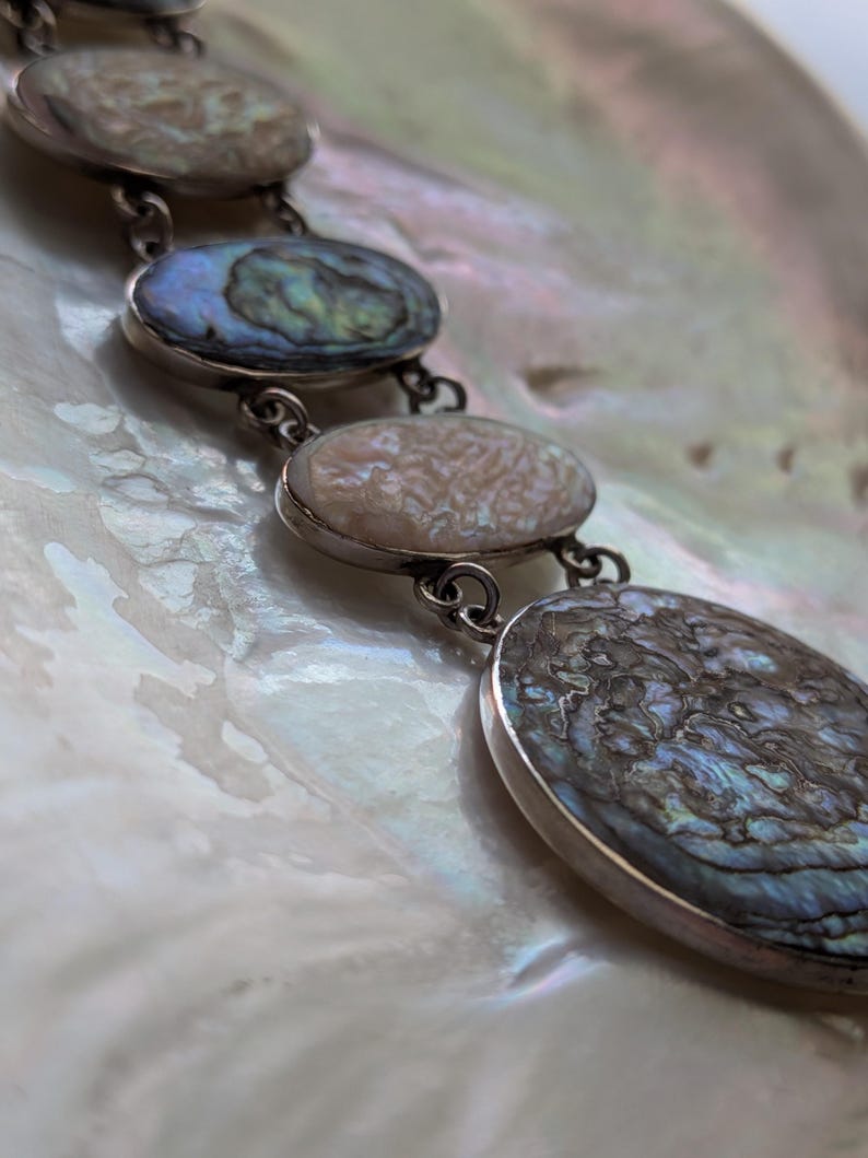 May include: A silver chain necklace with oval-shaped abalone shell pendants. The pendants have a iridescent blue, green, and brown color.