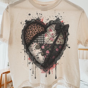 May include: A cream-coloured t-shirt with a large heart design. The heart is a patchwork of patterns, including leopard print, plaid, and floral designs, with a black lace border and a black bow. The design has a distressed, vintage aesthetic.