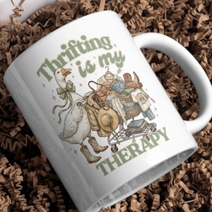 May include: White ceramic mug with the text "Thrifting is my Therapy" in green. The mug features a cartoon goose wearing a bow tie and cowboy boots, pushing a shopping cart filled with clothes and accessories.
