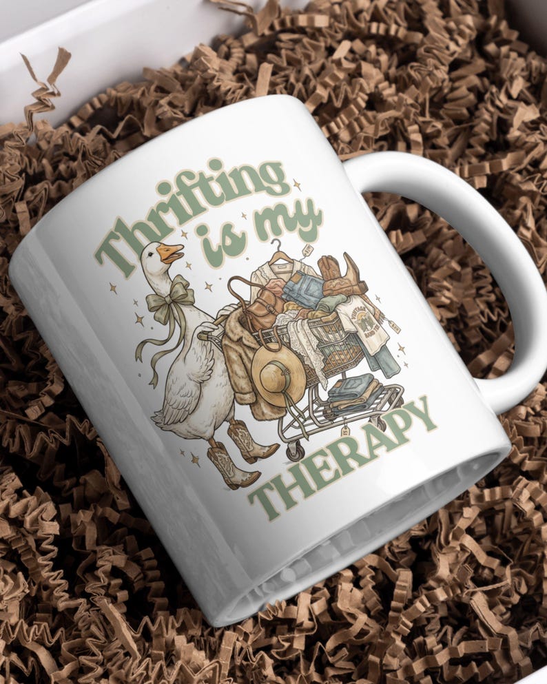 May include: White ceramic mug with the text "Thrifting is my Therapy" in green. The mug features a cartoon goose wearing a bow tie and cowboy boots, pushing a shopping cart filled with clothes and accessories.