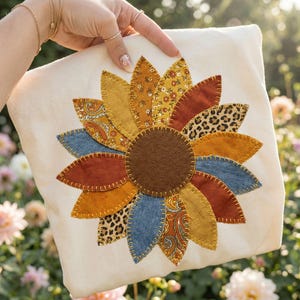 Pode incluir: Almofada creme com um grande desenho de girassol aplicado. As pétalas são feitas de tecidos de ganga, paisley e estampado de leopardo em tons de amarelo, laranja e azul. O centro é um círculo de feltro castanho.