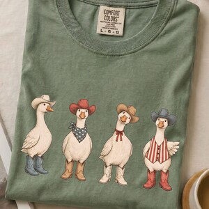 May include: A sage green t-shirt featuring a graphic of four white geese dressed as cowboys. Each goose wears a different colored cowboy hat and boots, with accessories including a star-patterned bandana, a red bow, and a red and white striped vest.