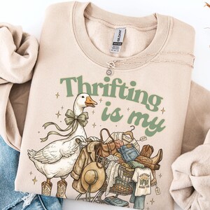 May include: Beige sweatshirt with the text "Thrifting is my" in green. A cartoon goose wearing cowboy boots and a bow is pushing a shopping trolley filled with clothes, a hat, and boots. A necklace is draped on the sweatshirt.