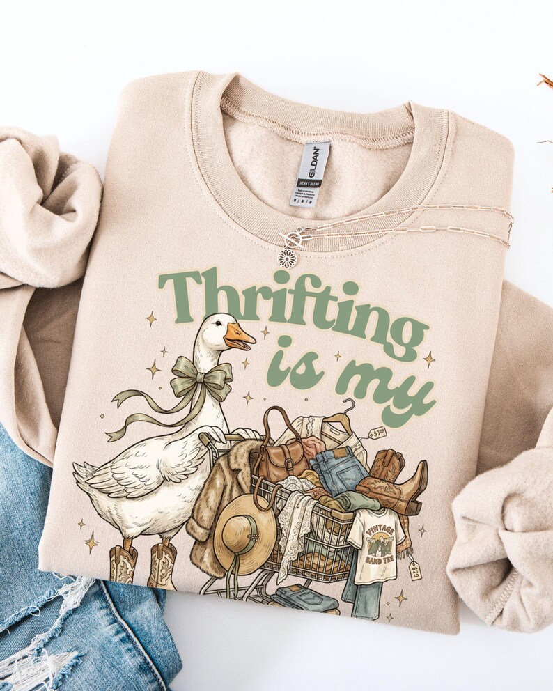 May include: Beige sweatshirt with the text "Thrifting is my" in green. A cartoon goose wearing cowboy boots and a bow is pushing a shopping trolley filled with clothes, a hat, and boots. A necklace is draped on the sweatshirt.