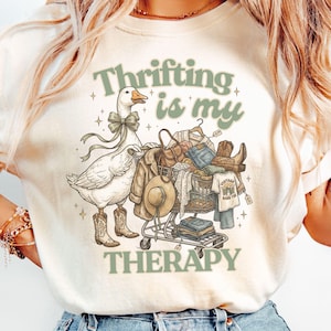 以下が含まれることがあります： 「Thrifting is my Therapy」の文字が緑色でプリントされたクリーム色のTシャツ。カウボーイブーツとリボンをつけたガチョウが、服やアクセサリーでいっぱいのショッピングカートを押しているグラフィック。