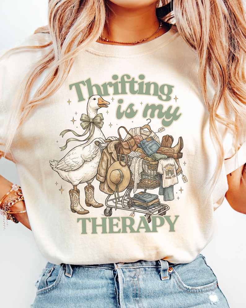 May include: A cream-coloured t-shirt with the text "Thrifting is my Therapy" in green. The graphic shows a goose in cowboy boots and a bow, pushing a shopping trolley filled with clothes and accessories.