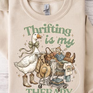 May include: Beige sweatshirt featuring a goose in cowboy boots and a bow tie, pushing a shopping trolley full of clothes. Text reads "Thrifting is my Therapy."