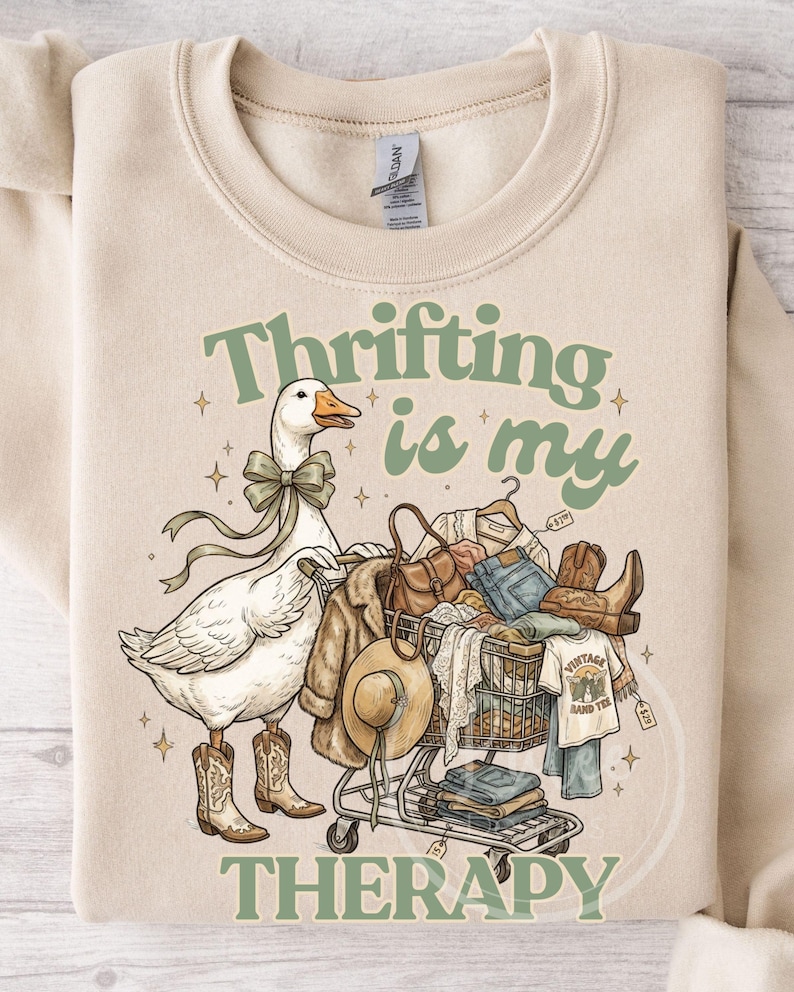 May include: Beige sweatshirt featuring a goose in cowboy boots and a bow tie, pushing a shopping trolley full of clothes. Text reads "Thrifting is my Therapy."