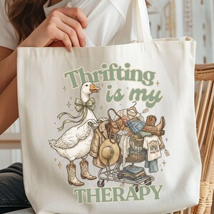 May include: Cream tote bag with a quirky illustration. A goose in cowboy boots and a bow tie stands next to a shopping trolley filled with clothes and a hat. The text "Thrifting is my therapy" is printed in green.