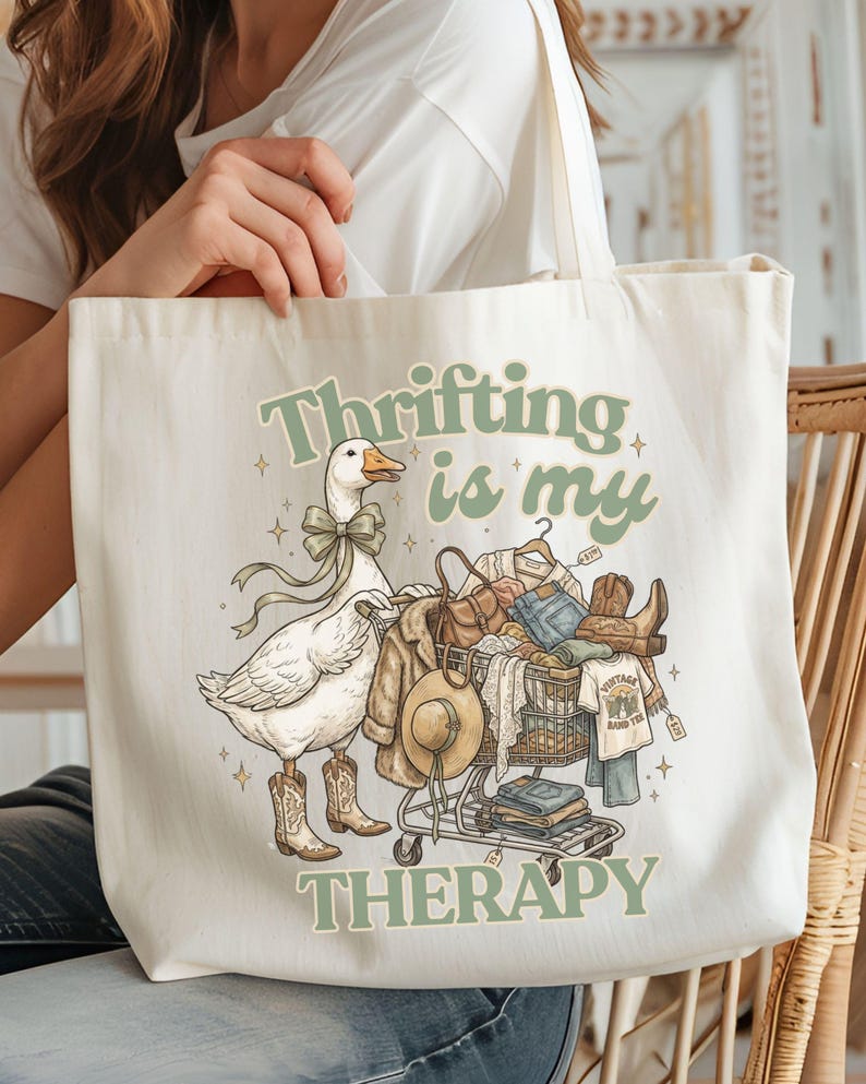 May include: Cream tote bag with a quirky illustration. A goose in cowboy boots and a bow tie stands next to a shopping trolley filled with clothes and a hat. The text "Thrifting is my therapy" is printed in green.
