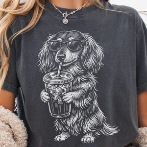 Puede incluir: Camiseta gris oscuro con una ilustración blanca de un perro salchicha con gafas de sol y sosteniendo una bebida helada. El perro está representado con pelo largo y una pose relajada. La camiseta es de estilo casual.