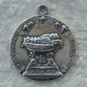 Peut inclure: Un pendentif en argent représentant l'enfant Jésus dans une crèche. Le pendentif est rond et porte les inscriptions "SS. VIRGEN" et "PIRA".