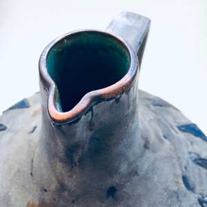 May include: A gray ceramic pitcher with a green glaze inside and a brown rim. The pitcher has a rustic, aged look.