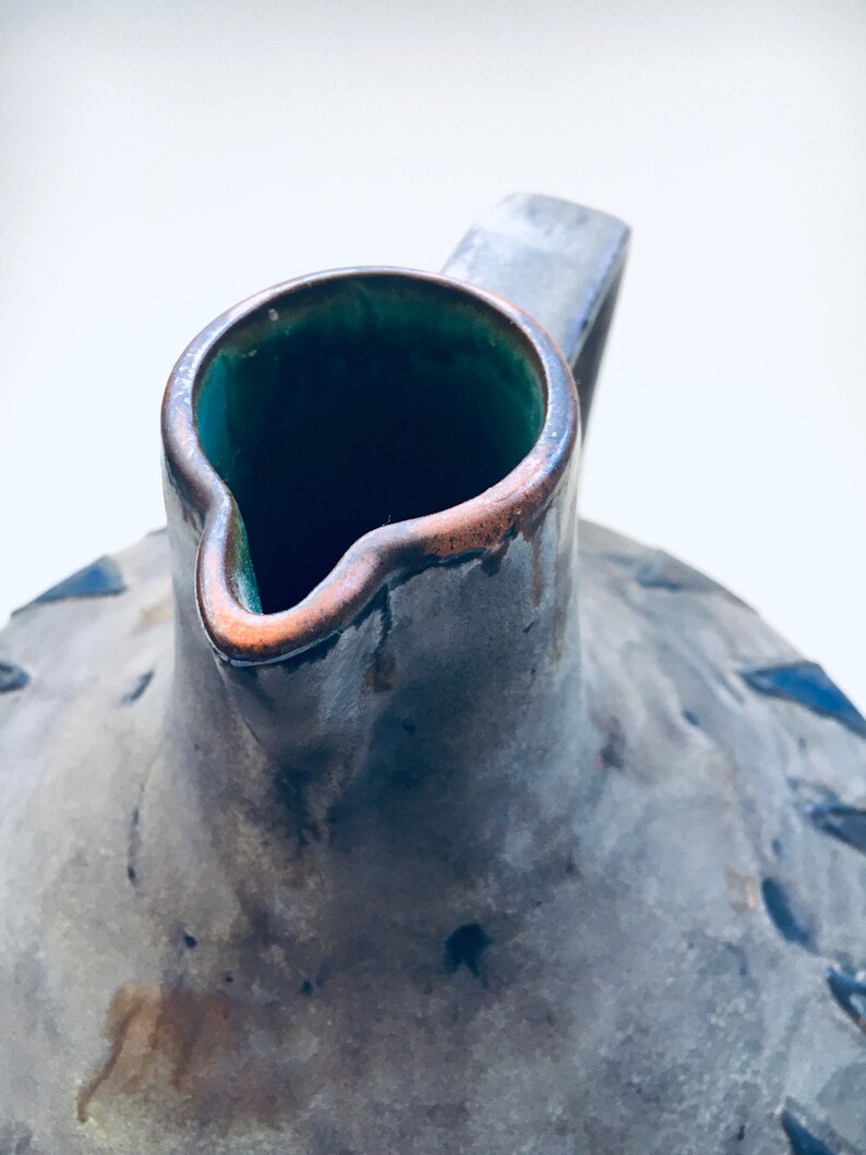 May include: A gray ceramic pitcher with a green glaze inside and a brown rim. The pitcher has a rustic, aged look.