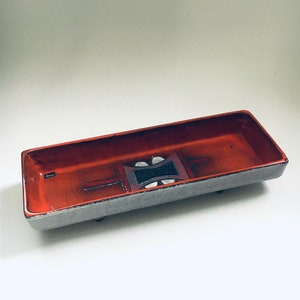 Könnte beinhalten: Eine rechteckige Servierplatte aus Keramik mit einem geometrischen Design in Rot-, Schwarz-, Weiß- und Grautönen. Die Platte hat einen erhöhten Rand und drei kleine Füße.