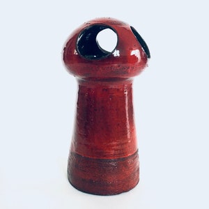 Puede incluir: Un jarrón de cerámica rojo con una parte superior en forma de hongo y varios agujeros. El jarrón tiene un acabado brillante y una base texturizada.