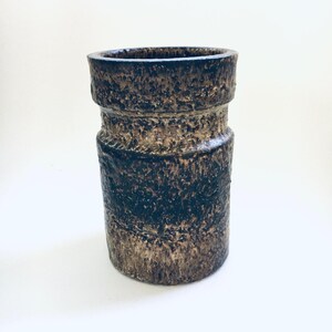 May include: A textured, cylindrical vase in shades of brown and black. The ceramic vessel has a rough, uneven surface with horizontal bands. The top rim is slightly flared, and the overall design suggests a vintage or rustic aesthetic.