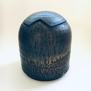Könnte beinhalten: Ein dunkelblauer Keramikbehälter mit Deckel. Der Behälter hat eine strukturierte Oberfläche mit einem Farbverlauf aus Braun- und Goldtönen. Der Deckel hat ein Zickzack-Design und eine dunklere blaue Farbe.