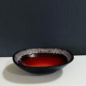 Może przedstawiać: Ręcznie robiona, owalna ceramiczna miska z czarnym, teksturowanym zewnętrzem. Wnętrze ma gradient kolorów, przechodzący od żywej pomarańczy u podstawy do głębokiej czerwieni i białej, wzorzystej krawędzi.