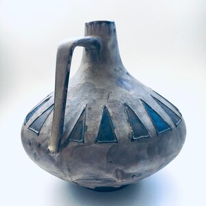 May include: A gray ceramic vase with a blue and gray geometric design. The vase has a wide base and a narrow neck with a handle.