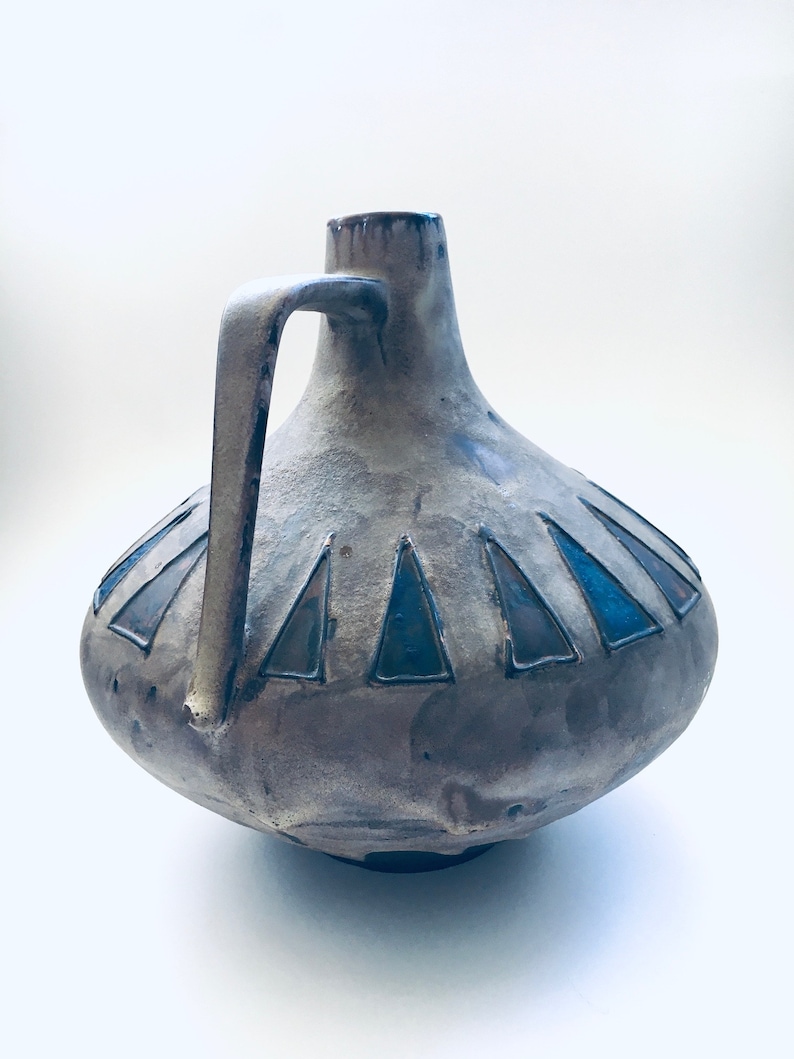 May include: A gray ceramic vase with a blue and gray geometric design. The vase has a wide base and a narrow neck with a handle.