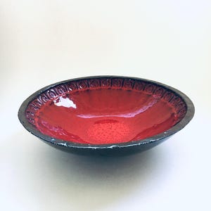Op de afbeelding: Een keramische kom met een glanzend rood interieur en een donkere, getextureerde buitenkant. De rand heeft een herhalend patroon van kleine, cirkelvormige ontwerpen. De kom heeft een ovale vorm en staat op een wit oppervlak.