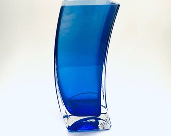 Könnte beinhalten: Eine kobaltblaue Glasvase mit geschwungenem Design. Die Vase hat einen quadratischen Boden und eine abgerundete Oberseite.