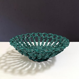 May include: A green ceramic basket with a woven design. The basket is oval-shaped and sits on a small base. The basket's intricate pattern creates a shadow on the white surface below. The background is a mix of black and white.