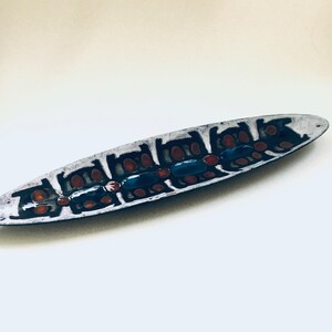 May include: A long, white ceramic serving dish with a geometric pattern in black, red, and blue. The dish is oval shaped and has a glossy finish.