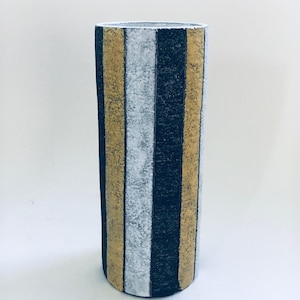 Puede incluir: Un jarrón alto y cilíndrico con rayas verticales doradas, plateadas y azul marino. El jarrón tiene una superficie texturizada y un diseño sencillo y moderno. El jarrón es adecuado para exhibir flores o como elemento decorativo.