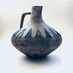 May include: A brown ceramic pitcher with a wide base and a narrow neck. The pitcher has a handle and a geometric design of blue and orange triangles around the middle.