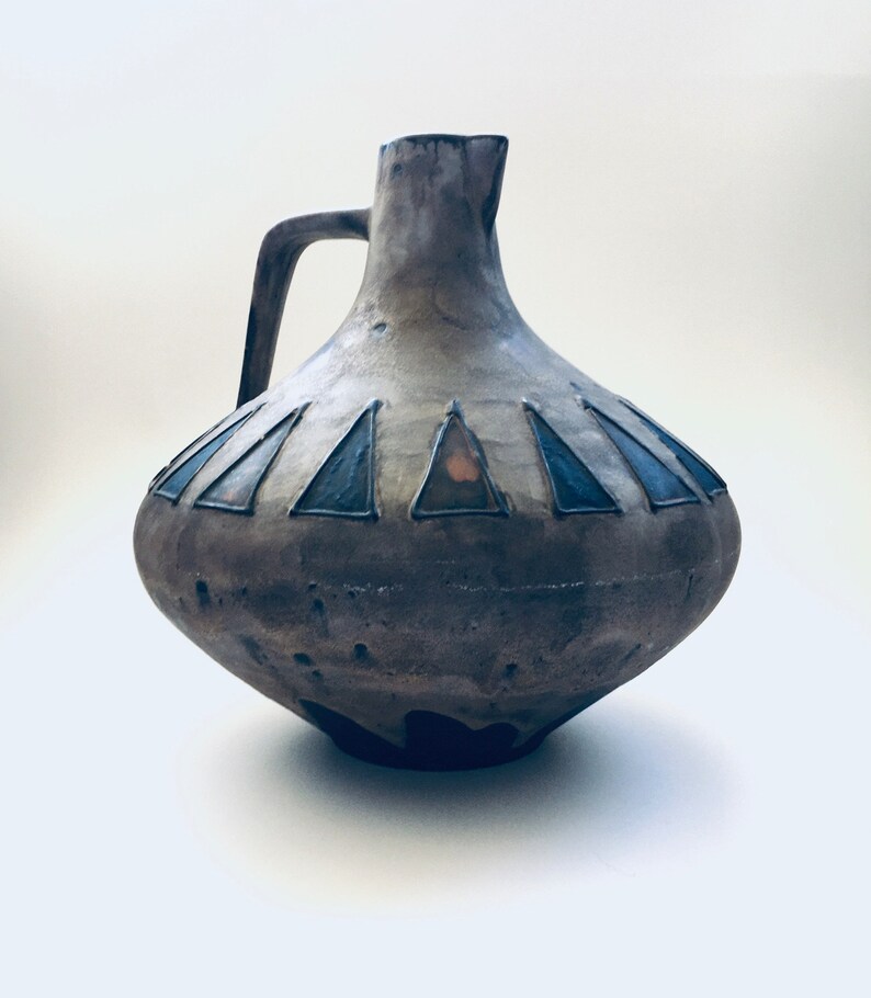 May include: A brown ceramic pitcher with a wide base and a narrow neck. The pitcher has a handle and a geometric design of blue and orange triangles around the middle.