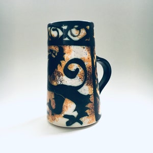 Puede incluir: Una taza de cerámica con un diseño negro y marrón que presenta un león estilizado y motivos florales. La taza tiene un asa y una base redondeada.