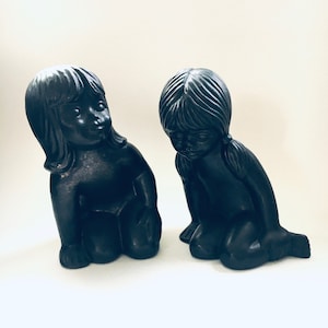 Puede incluir: Dos figuritas de cerámica negra de niñas con el pelo largo, ambas arrodilladas con la cabeza gacha. La niña de la izquierda mira hacia la derecha, mientras que la niña de la derecha mira hacia abajo.